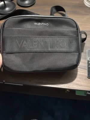 Svart axelväska från Valentino - Snygg svart axelväska från Valentino med justerbar axelrem och diskret logga både på väskan och remmen. Väskan har en rektangulär form och är tillverkad i slitstarkt tygmaterial. Perfekt för att bära dina viktigaste grejer med stil.