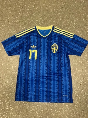 Blå Sverige fotbollströja Adidas - Säljer en blå fotbollströja från Adidas med Sveriges landslagsemblem och nummer 17 i gult. Tröjan har gula detaljer, tre ränder på axlarna och namntryck 'Gyökeres' på ryggen. Mönstrad design och korta ärmar, perfekt för match eller träning.
