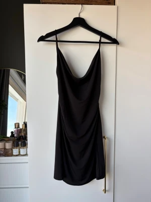 Svart kort klänning med tunna band - Snygg svart kort klänning med tunna axelband och draperad urringning. Klänningen har en enkel och stilren design som ger en elegant look. Tillverkad i ett mjukt och följsamt material som faller fint längs kroppen.