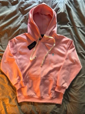 Rosa hoodie från Polo Ralph Lauren - Snygg rosa hoodie från Polo Ralph Lauren med klassisk huva och dragsnören. Har en broderad logga i ljusblått på bröstet och en stor magficka. Mjuk bomullskänsla och ribbade muddar vid ärmslut och nederkant. Perfekt för en chill och trendig look.