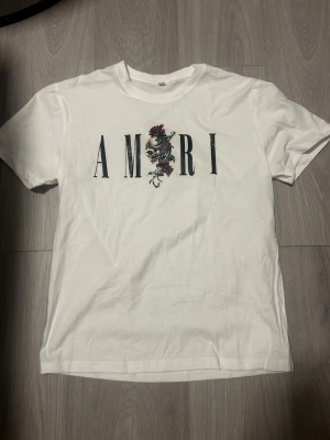 Vit AMIRI t-shirt med tryck - Vit t-shirt från AMIRI med stort tryck framtill. Trycket består av AMIRI-loggan med en döskalle, rosor och revolver i mitten. Klassisk passform och rund halsringning. Perfekt för dig som gillar streetwear med edge. Storleken är M o T-shirten är använd ett par gånger men visar inga tecken på det förut att det finns nån prick vid kragen som går bort i tvätten. 
