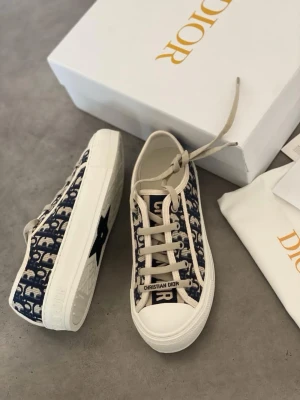Dior Oblique sneakers i blått och vitt - Snygga Dior Oblique sneakers med blått och vitt monogrammönster, vita snören och robust vit sula. Klassisk låg modell med tydlig Dior-logga på sidan och tungan. Perfekta för dig som vill sticka ut med exklusiv streetstyle.