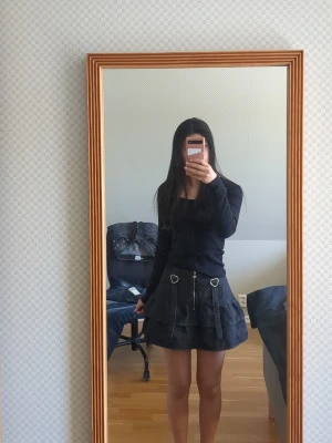 Svart kortkjol med hjärtformade detaljer - Svart kortkjol med volanger och två hjärtformade spännen framtill. Kjolen har dragkedja i mitten och dekorativa band. Perfekt för dig som gillar en edgy och söt stil. Pris går självklart att diskutera och säljer för att den inte kommer till användning. 