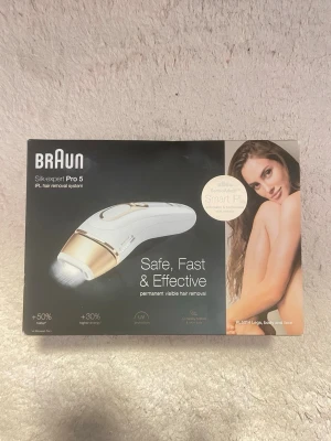 Braun Silk-expert Pro 5 IPL hårborttagare - IPL hårborttagningssystem från Braun, modell Silk-expert Pro 5. Enheten är vit med gulddetaljer och används för permanent synlig hårborttagning på ben, kropp och ansikte. Utrustad med Smart IPL-teknik och UV-skydd.