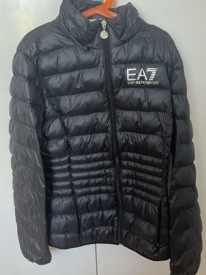 Svart pufferjacka från Emporio Armani - Svart pufferjacka från Emporio Armani EA7 med hög krage och dragkedja framtill. Jackan har ett stilrent, quiltat mönster och logga på bröstet. Perfekt för kalla dagar och ger en sportig, trendig look. Materialet är lätt och isolerande.