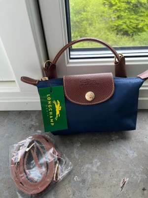 Blå handväska  - Stilren blå handväska  med bruna läderdetaljer och guldfärgad knapp. Väskan har ett kort handtag och medföljande justerbar axelrem i brunt läder. Materialet är nylon och läder, perfekt för dig som gillar klassisk design.