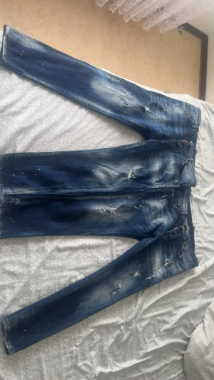 Blå  jeans från Dsquared2 - Säljer ett par blå jeans från Dsquared2 med fina , färgstänk och distressed detaljer. Jeansen har klassisk femficksmodell, knappgylf och smal passform. Perfekta för dig som gillar en edgy och trendig stil.