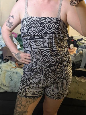 Svartvit mönstrad jumpsuit - Snygg svartvit jumpsuit med grafiskt mönster och smala axelband. Modellen har resår i midjan för en bekväm passform och korta ben. Perfekt för en trendig och avslappnad look. Materialet känns lätt och luftigt, perfekt för varma dagar.