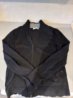 Svart ziptröja från Pier One - Svart ziptröja från Pier One med ribbad ståkrage och muddar vid ärmslut och nederkant. Tröjan har hel dragkedja framtill och är tillverkad i ett mjukt material, perfekt för en avslappnad stil.