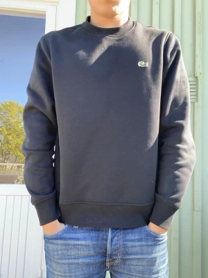 Svart sweatshirt från Lacoste - Klassisk svart sweatshirt från Lacoste med rund halsringning och diskret broderad logotyp på bröstet. Tröjan har ribbade muddar vid ärmslut och nederkant. Perfekt för en avslappnad och stilren look.