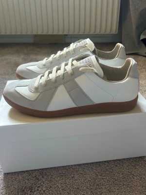 Maison margiela gats  - Säljer ett par stilrena vita sneakers med beige mockadetaljer och bruna gummisulor. Skorna har klassisk låg profil, vita snören och paneler i både läder och mocka. Perfekta för dig som gillar minimalistisk och clean stil.