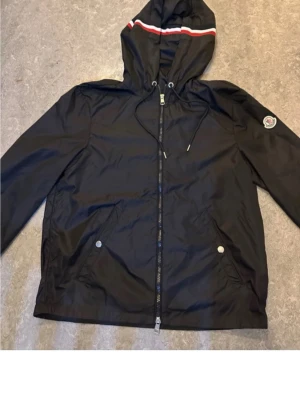 Moncler windbreaker - Svart vindjacka från Moncler med huva och dragsko. Jackan har dragkedja framtill, två fickor med tryckknappar och Moncler-logga på vänster ärm. Huvan har en vit och röd rand som snygg detalj. Tillverkad i lätt och smidigt material som står emot vind. Modellen är 184 75kg