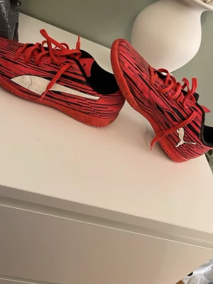 Röda Puma fotbollsskor strl 39 - Säljer ett par röda fotbollsskor från Puma med svarta och vita detaljer samt snörning. Skorna har ett sportigt mönster med svarta ränder och den klassiska Puma-loggan på sidan. Yttermaterialet är syntet och insidan är svart. Perfekta för dig som vill sticka ut på planen.