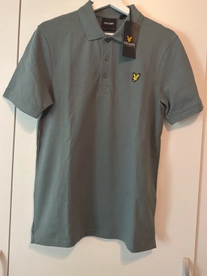Grön pikétröja från Lyle & Scott - Snygg grå pikétröja från Lyle & Scott med klassisk krage, knappar framtill och den ikoniska gula loggan på bröstet. Tillverkad i mjuk bomull och har korta ärmar. Perfekt för en clean och stilren look.