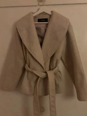 Beige kappa från Vero Moda med skärp - Stilren beige kappa från Vero Moda med bred krage och knytskärp i midjan. Kappan har en klassisk, rak passform och är tillverkad i ett mjukt tyg som ger en snygg siluett. Perfekt för dig som gillar minimalistisk och trendig stil.