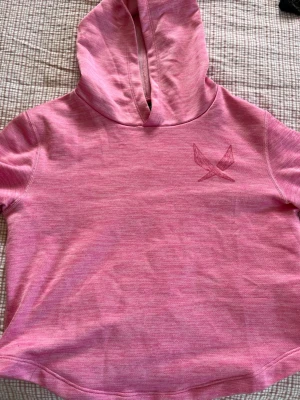 Rosa hoodie med kort ärm och luva - En söt rosa hoodie med korta ärmar och huva. Den har en tryckt logga på bröstet och är gjord i ett mjukt, stretchigt material. Perfekt för en avslappnad och sportig stil. Passar dig som gillar färg och vill sticka ut lite extra. Från soc och i storlek 146/152