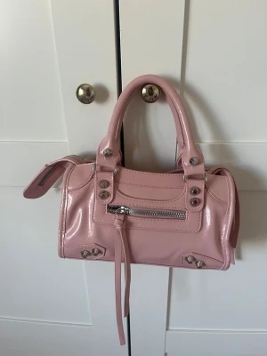 Ljusrosa handväska med nitar - Söt ljusrosa handväska i glansigt material med silverfärgade nitar och dragkedjedetaljer. Väskan har två handtag och en liten ficka framtill med dragkedja och dekorativa remmar. Perfekt för dig som gillar trendiga accessoarer.