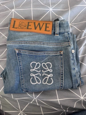 LOEWE NICHE JEANS  - Snygga blå jeans från Loewe med klassisk femficksmodell. Baksidan har en stor Loewe-logga broderad i vitt på ena bakfickan och ett stort läderpatch med Loewe-tryck i midjan. Jeansen är raka i modellen och tillverkade i denim.
