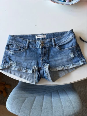 Blå lågmidjade jeansshorts från Tally Weijl - Snygga blå lågmidjade jeansshorts från Tally Weijl med slitna detaljer och fransig kant. Klassisk femficksmodell med låg midja och dubbla knappar framtill. 