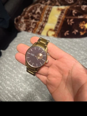 Guldfärgad klocka från Axcent - Stilren guldfärgad klocka från Axcent med metallarmband och mörk urtavla. Klockan har minimalistisk design med tydliga markeringar och ett klassiskt spänne. Perfekt accessoar för dig som gillar en clean look.