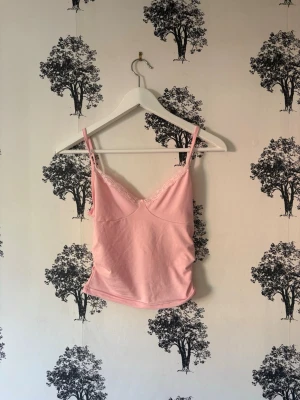 Rosa linnetopp med spetsdetalj - Söt ljusrosa linnetopp med tunna axelband och spetskant vid urringningen. Toppen har en feminin v-ringad form och är croppad i modellen. Perfekt att styla med jeans eller kjol för en trendig look.