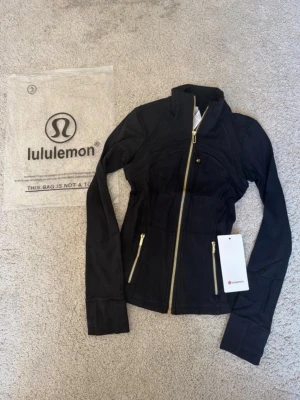 Svart träningsjacka från Lululemon black gold svart guld S stl 2  - Snygg svart träningsjacka från Lululemon med guldiga dragkedjor och figurnära passform. Jackan har hög krage, långa ärmar och två fickor framtill med dragkedja. Perfekt för gymmet eller löprundan. Storlek 2 passar S 