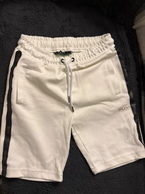 Vita BOSS shorts med svarta detaljer - Snygga vita shorts från BOSS med svarta sidopaneler och logga på benet. De har elastisk midja med snörning, två fickor fram och en bakficka. Perfekta för en avslappnad och trendig stil.