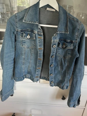Klassisk blå jeansjacka - Snygg blå jeansjacka med två bröstfickor med lock och knapp, samt dekorativa sömmar. Jackan har en rak passform, krage och knäppning framtill med metallknappar. Perfekt att slänga över en t-shirt för en avslappnad look.
