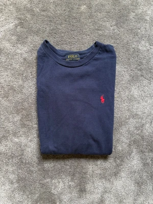 Mörkblå t-shirt från Polo Ralph Lauren - Klassisk mörkblå t-shirt från Polo Ralph Lauren med röd broderad logga på bröstet. T-shirten har rund halsringning och är tillverkad i mjuk bomull. Perfekt för en stilren och enkel look.