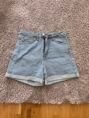 Ljusblå high waist jeansshorts Zara - Säljer ett par ljusblå denimshorts från Zara med hög midja och uppvikta benslut. Klassisk femficksmodell med knapp och dragkedja framtill samt läderpatch bak i midjan. Perfekta till sommaren och lätta att matcha med olika stilar.