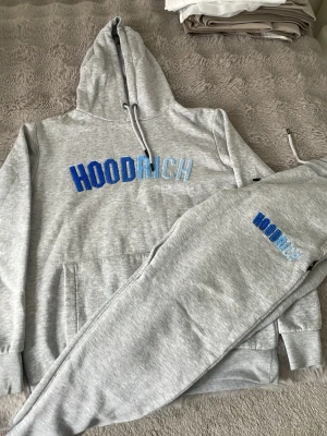 Grå Hoodrich hoodie och byxor set - Säljer ett grått mjukisset från Hoodrich med blå och ljusblå broderad logga på både hoodie och byxor. Hoodien har huva med dragsko och magficka, byxorna har resår i midjan och vid benslut. Perfekt för en chill och stilren look.