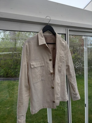 Beige overshirt i linne - Stilren beige overshirt i linne med klassisk krage och bröstfickor med lock. Knäpps med bruna knappar framtill och har raka ärmar. Perfekt lager på lager-plagg för en avslappnad look.