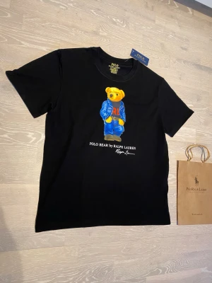 Svart Polo Bear t-shirt Ralph Lauren - Svart t-shirt från Ralph Lauren med ikoniskt Polo Bear-tryck på bröstet. Björnen har blå jacka och jeans, samt röd RL-logga på tröjan. Storleken är M.      Priset kan diskuteras och jag skickar inom 24 Tim☺️💨