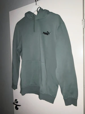 Grön hoodie från Puma med logga - Snygg grön hoodie från Puma med svart logga på bröstet. Klassisk modell med huva och dragsko samt stor magficka framtill. Perfekt för en avslappnad och sportig stil. Mjuk och skön bomullsblandning som passar året runt.