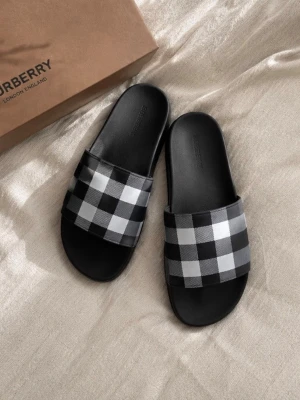 Burberry tofflor  - Säljer ett par snygga slides från Burberry med klassiskt svartvitt rutmönster. Sandalerna har en bred rem i textil med Burberrys ikoniska mönster och en svart sula i syntetmaterial. Perfekta för en trendig och avslappnad stil. Box och dustbag följer med 