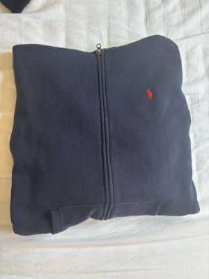Mörkblå hoodie från Polo Ralph Lauren - Mörkblå hoodie från Polo Ralph Lauren med dragkedja framtill och den klassiska röda loggan broderad på bröstet. Tillverkad i mjukt bomullsmaterial som känns skönt mot huden. Perfekt för en avslappnad och stilren look.