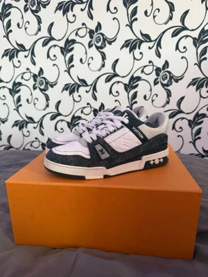 Louis Vuitton vita och svarta sneakers - Snygga Louis Vuitton sneakers i vitt och svart med klassisk monogramdetalj. Skorna har låga skaft, vita snören och robust sula med LV-logga undertill. Materialet är en mix av läder och textil för en exklusiv känsla och cool look.