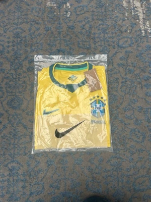 Gul Brasilien Nike fotbollströja - Säljer en gul Brasilien fotbollströja från Nike med gröna detaljer och CBF-emblem på bröstet. Tröjan har korta ärmar, rund halsringning med grönt kantband och Nike-logga i grönt. Perfekt för dig som älskar fotboll och vill stötta Brasilien.