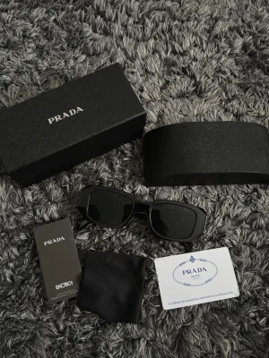 Svarta solglasögon från Prada - Snygga svarta solglasögon från Prada med rektangulära bågar. Kommer med originalfodral, putsduk och äkthetskort. Glasögonen har en stilren och modern design som passar till alla tillfällen.