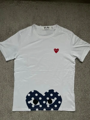 Vit Comme des Garçons Play t-shirt - Säljer en vit t-shirt från Comme des Garçons Play med det ikoniska röda hjärtat på bröstet och ett stort blått hjärta med vita prickar och ögon längst ner. Klassisk passform och korta ärmar, tillverkad i mjuk bomull.