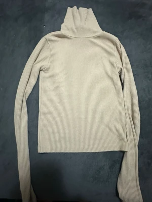 Beige ribbad polotröja - Säljer en beige ribbad polotröja med långa ärmar och hög krage. Tröjan har en slim fit och är perfekt för lager-på-lager-stil. Materialet är mjukt och stretchigt för en bekväm känsla.