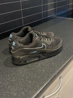 Nike Air Max 90 svarta sneakers - Säljer ett par svarta Nike Air Max 90 sneakers med klassisk siluett och synlig Air-dämpning i sulan. Skorna har vita detaljer på loggan och swoosh, samt svarta snören och ovandel i textil och syntet. Perfekta för dig som gillar streetwear och sportig stil.