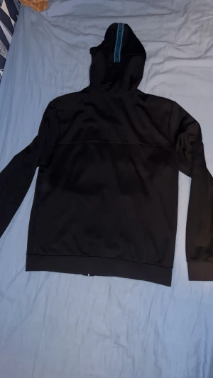 Svart hoodie från BOSS med blå detaljer - Snygg svart hoodie från BOSS med dragkedja och blå logga på bröstet. Huva med blå snören och blå textdetalj längs huvkanten. Tillverkad i mjukt material som känns skönt mot huden. Perfekt för en avslappnad och stilren look. OBS lite slitas vid armbågarna
