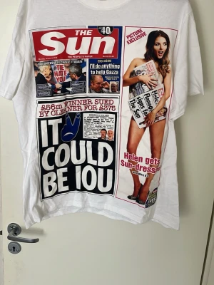 Vit t-shirt med The Sun tidningsomslag - Unik vit t-shirt med stort tryck av The Sun tidningsomslag på framsidan. Färgglad och iögonfallande design med text och bild i rött, svart, blått och vitt. Klassisk passform och rund hals. Perfekt för dig som gillar statement-plagg och popkultur.