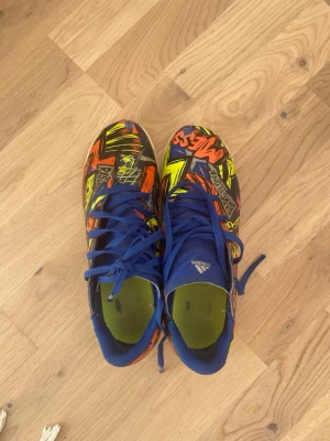 Adidas Messi färgglada fotbollsskor inomhus  - Säljer ett par Adidas Messi fotbollsskor med grymt färgstarkt mönster i blått, gult, orange och svart. Skorna har blå snörning och innerfoder, samt coola grafiska detaljer och Messi-tryck på ovandelen. Perfekta för dig som vill sticka ut på planen!