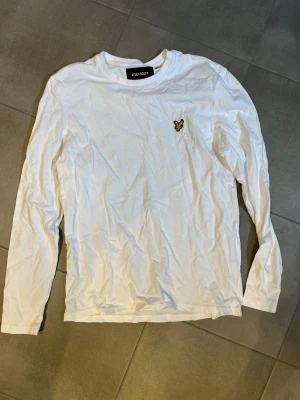 Vit långärmad t-shirt från Lyle & Scott - Vit långärmad t-shirt från Lyle & Scott med rund hals och klassisk gul logga på bröstet. Tillverkad i mjuk bomull för en skön känsla och enkel stil. Perfekt till jeans eller shorts för en clean look.