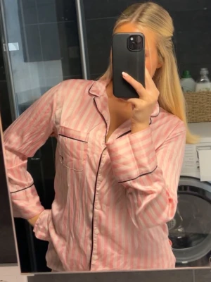 Rosa randig pyjamas från Victorias Secret - Säljer en jättesöt pyjamas i rosa och vitt från Victoria Secret. Setet har långärmad skjorta med krage, bröstficka och svarta passpoaldetaljer samt matchande byxor. Materialet är superskönt och den är sparsamt använd. Den har en miniliten svart prick ovanför fickan, se sista bilden 🩷🩷