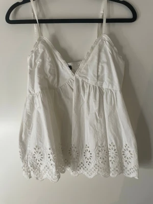 Vit broderad linnetopp med spets - Söt vit linnetopp med tunna axelband, v-ringning och spetsdetaljer längs kanterna. Toppen har broderade blommor och hålmönster längst ner samt en luftig passform. Perfekt för varma dagar och somriga outfits.