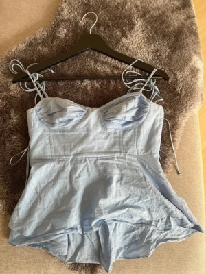 Snygg topp från Zara - Supersöt ljusblå linnetopp med tunna axelband som knyts på axlarna. Toppen har en lätt utsvängd passform och markerade sömmar vid bysten för en snygg siluett. Perfekt för varma dagar och enkel att styla med jeans eller kjol.