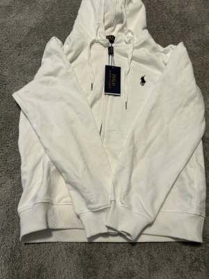 Vit hoodie från Polo Ralph Lauren - Klassisk vit hoodie från Polo Ralph Lauren med dragkedja framtill och justerbar huva med snören. Broderad logga i svart på bröstet och ribbade muddar vid ärmar och nederkant. Tillverkad i mjuk bomull för en skön och avslappnad look.